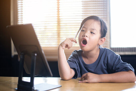 Curious young Asian girl thinking and talking during online class with tablet, little child learning language or practicing speech with digital device at home.