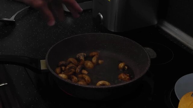Chef saut&eacute;es mushrooms in a non-stick pan on a stovetop. Culinary process of cooking sliced button mushrooms with soy sauce. Healthy homemade meal preparation.