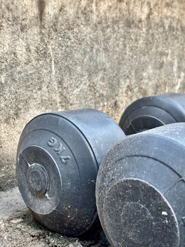 Two black 2kg dumbbells resting on a dirty ground against a rough concrete wall. Gritty outdoor home gym equipment, physical fitness and weight training concept.