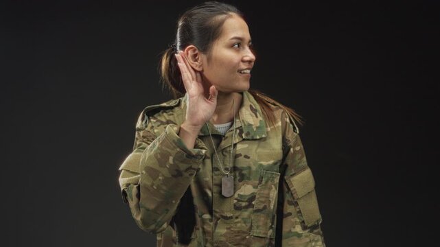 Woman soldier in camouflage uniform with dogtags holds hand to ear in studio listening pose; duty readiness.