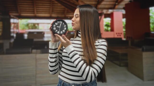 Woman in striped turtleneck holding a black clock and puckering lips while presenting it in a building; playful affection.