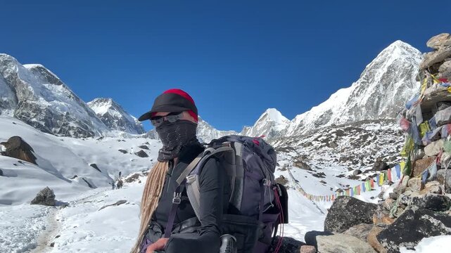Female trekker with backpack, dreadlocks and face covered records selfie video near memorial on Everest Base Camp trail, snowy Himalayan mountains, high altitude around 5000m, Lobuche route