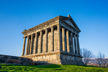 Fototapeta premium Sunlit ancient Garni temple with stone columns, grassy hill, and bare trees