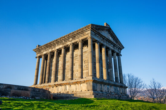 Sunlit ancient Garni temple with stone columns, grassy hill, and bare trees