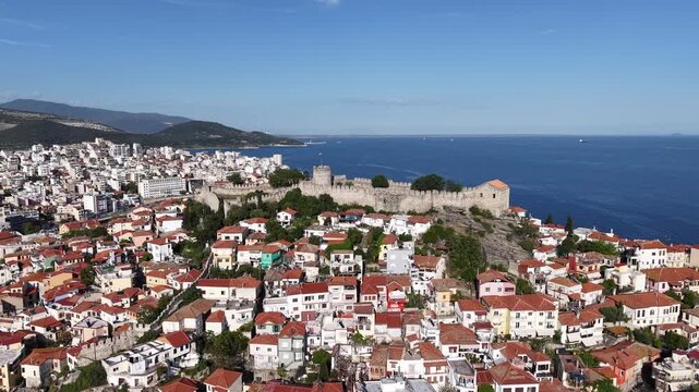 Aerial shot of tightly packed red-roof houses with fortified stone walls and blue water in the background.