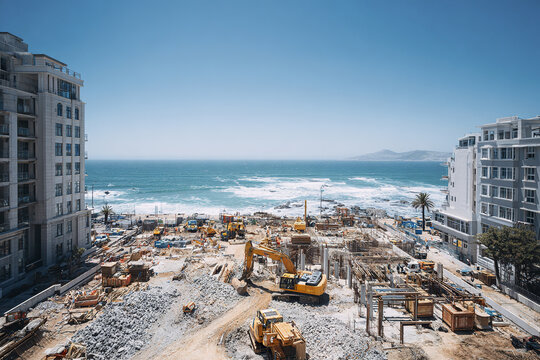 Urban construction site with heavy machinery and building materials near ocean shore under clear blue sky, showing active development and coastal landscape view