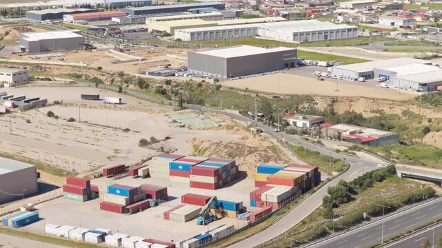 Aerial drone view of modern logistics warehouses situated alongside a major highway, featuring vibrant stacks of multicolored shipping containers and a sprawling industrial zone in the background. 