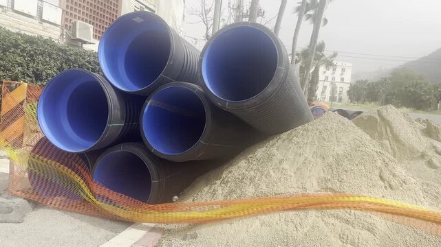 Corrugated plastic construction pipes stacked in a pyramid on a construction site, near a pile of sand and fenced with red safety mesh, beginning of construction