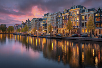 Fototapeta premium Canal with traditional narrow houses reflecting in water during colorful sunset sky, autumn trees lining street, peaceful urban scene with parked cars and warm lights