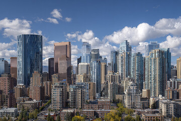Obraz premium Modern cityscape skyline with tall glass and concrete buildings under bright blue sky with scattered white clouds, showcasing urban architecture and vibrant city life