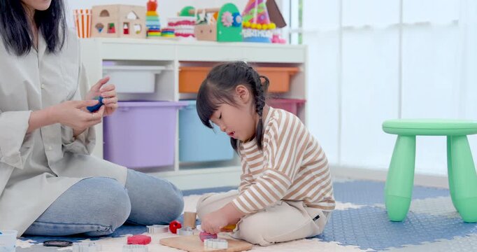 Asian preschool girl sits beside single mother in bright playroom, clapping during playful learning routine as small toy pieces rest nearby, building rhythm, attention, confidence, warm family bond.
