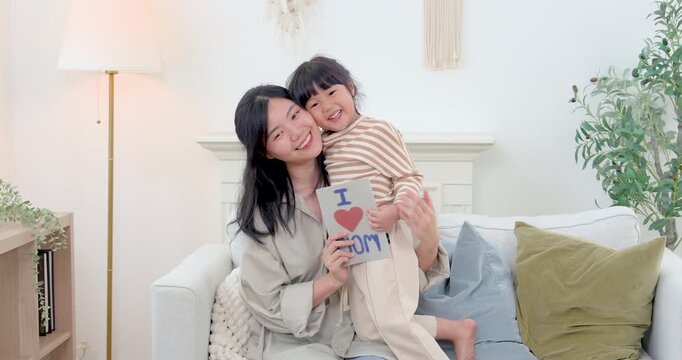 Asian single mother poses with smiling preschool daughter holding handmade Mothers Day card near sofa, sharing proud family bonding, gratitude, nurturing childcare care, joyful home celebration.
