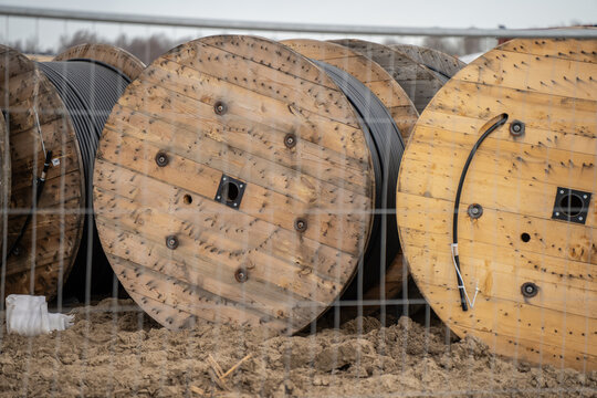 Large wooden cable reels behind wire fence on sandy construction site with black utility cable for electrical infrastructure project logistics