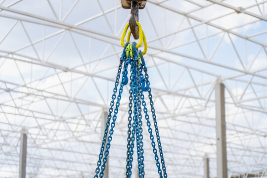 Blue chain sling assembly hanging from crane hook below steel roof framework for structural lifting and industrial facility construction tasks