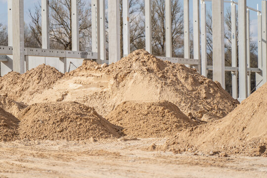 Large piles of sand and aggregate staged on warehouse construction site beside concrete column grid for earthworks, grading, and material supply use