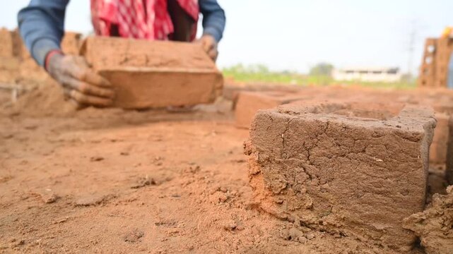 A person manually shaping bricks in an open field. Using a rectangular wooden mold and raw, reddish-brown clay, the worker skillfully forms each brick on the dusty ground. Slow motion video.
