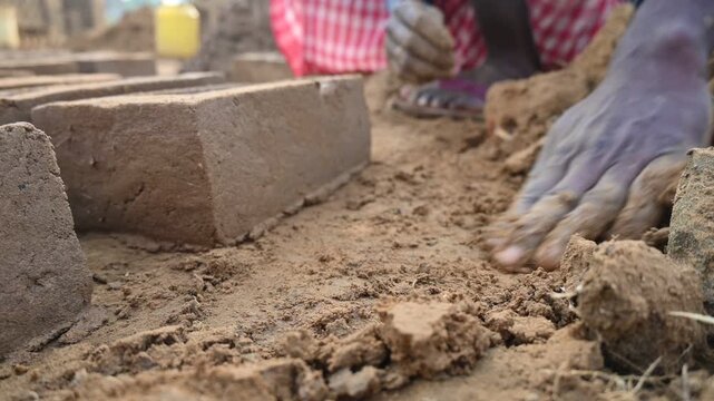A person manually shaping bricks in an open field. Using a rectangular wooden mold and raw, reddish-brown clay, the worker skillfully forms each brick on the dusty ground. Slow motion video.