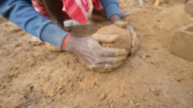 A person manually shaping bricks in an open field. Using a rectangular wooden mold and raw, reddish-brown clay, the worker skillfully forms each brick on the dusty ground. Slow motion video.