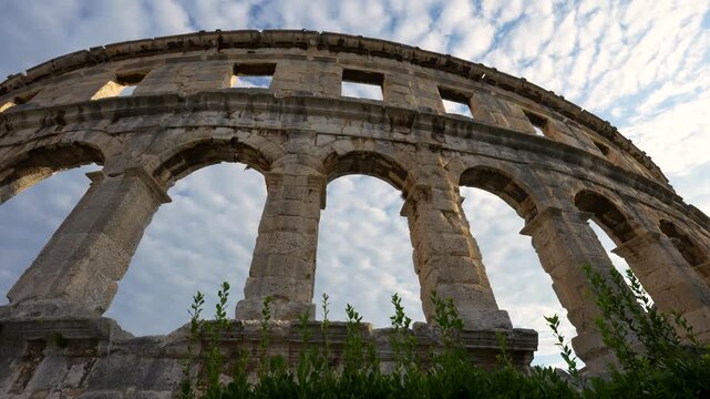 Pula, Croatia - August 18, 2025: The Pula Arena stands as a monumental ancient Roman amphitheater, its towering limestone arches reaching toward a soft, cloud-filled sky in the late afternoon.