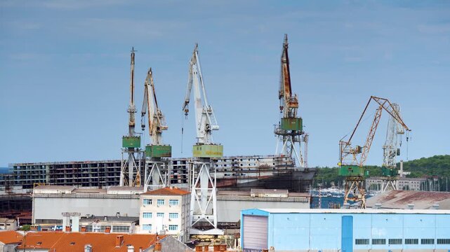 Pula, Croatia - August 18, 2025: Industrial harbor cranes stand tall over a shipyard in Pula, with unfinished vessel hulls and coastal buildings under a wide, clear blue sky.