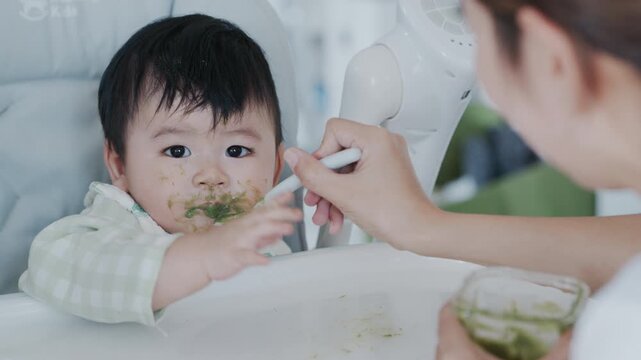 Happy Asian baby boy in high chair smiling at mother while being spoon-fed. Close-up view with natural light in a home