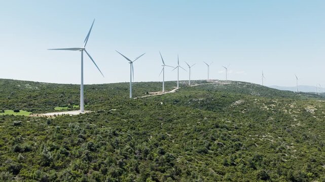 Multiple wind turbines with spinning blades standing in a row along a green hill, generating clean and sustainable electrical power from the wind against a clear blue sky on a sunny day