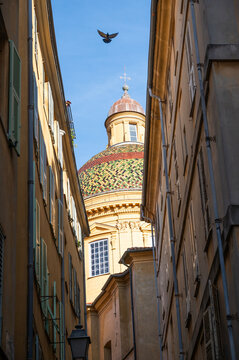 Narrow street view in the old town of Nice, France, featuring a colorful tiled dome church framed by traditional Mediterranean buildings. Warm sunlight and strong architectural perspective create a ty