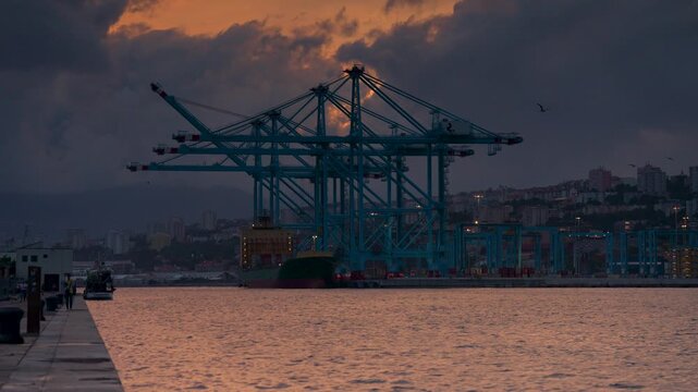 Rijeka, Croatia - August 21, 2025: Large blue gantry cranes tower over a container ship at a busy industrial port during a dramatic sunset. A coastal city climbs the hills in the background.