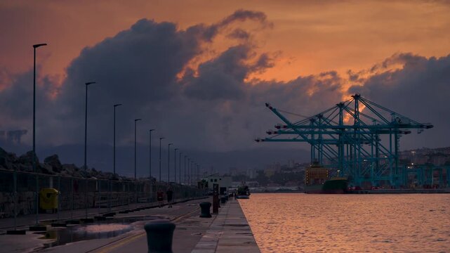 Rijeka, Croatia - August 21, 2025: The industrial silhouette of port cranes and a docked ship stands against a dramatic sunset sky in Rijeka, with a long stone pier stretching toward the calm water.