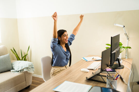 Latin woman celebrating achievement, working from home office, raising arms in excitement, enjoying professional victory