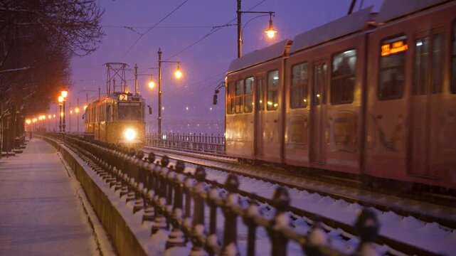 Budapest, Hungary - January 6, 2026: A yellow tram travels along snow-covered tracks beside the Danube in Budapest, illuminated by glowing streetlights against a purple winter twilight.