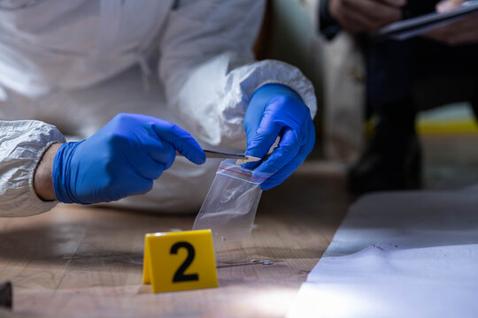Forensic specialist in protective suit and blue gloves collecting evidence with tweezers at a marked crime scene, assisting investigation