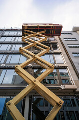 Naklejka premium Yellow scissor lift elevated against faceted geometric stone building facade under cloudy sky