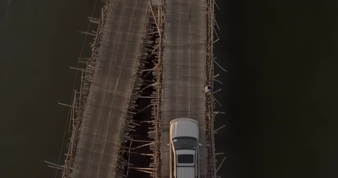 Backward top down aerial view of cars and motorcycles crossing parallel bamboo bridges at sunset.