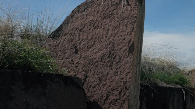 Handheld static close-up shot of the rough, reddish textured stone surface at the Great Salbyk Kurgan in Khakassia. The highly detailed ancient megalith stands amidst wild grass under a clear blue sky