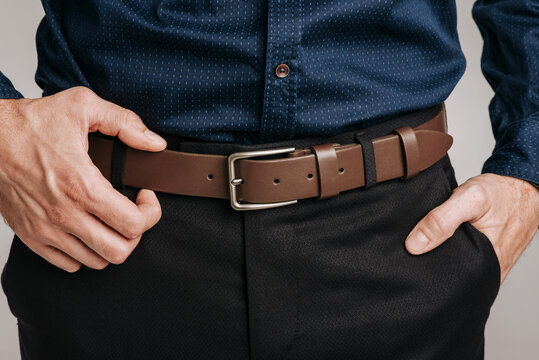 Close-up of a man wearing a classic brown leather belt with a metal buckle on dark trousers and a blue shirt.