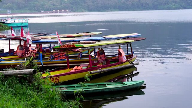 Panning shot of colorful wooden tourist boats equipped with life jackets, docked on the grassy bank of Selorejo Reservoir in Malang, Indonesia, waiting to transport visitors across the lake.