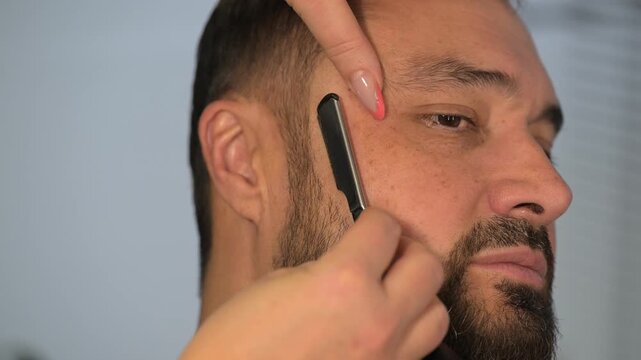 Barber trimming beard of adult man with straight razor in barbershop. Close-up of professional male grooming and beard care, hairstylist shaping beard during haircut service in modern salon.
