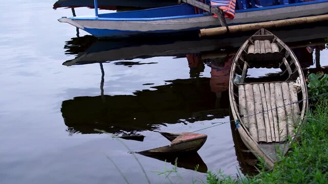 Traditional wooden sampan moored next to a broken, sunken boat wreck at the grassy edge of Selorejo Reservoir, Malang, Indonesia, illustrating themes of abandonment and rural decay.
