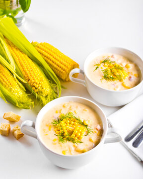 Two cups of creamy corn soup garnished with fresh dill and corn kernels, served with croutons and fresh corn cobs on a white table.