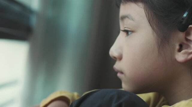 Close up side profile of sad Asian girl face looking away with selective focus. Lonely child sitting near window feeling bored, depressed, unhappy, thinking or waiting in solitude at home.