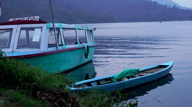 Small wooden fishing boat moored beside a tourist passenger vessel at the grassy edge of Selorejo Reservoir, Malang, Indonesia, during calm twilight.