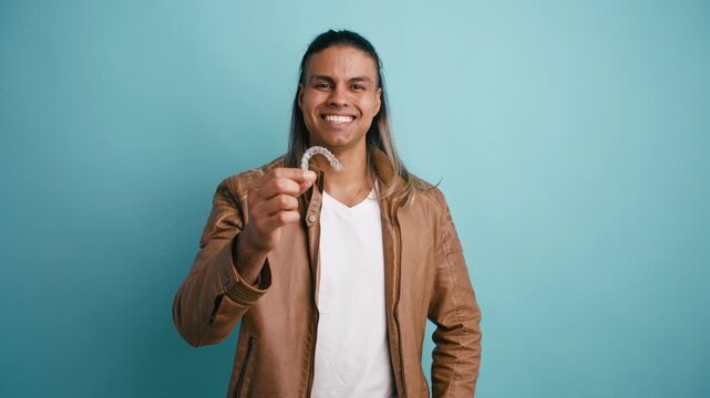 Happy Latino man with long hair showing a transparent dental aligner. Orthodontic treatment and removable straightener for a perfect smile