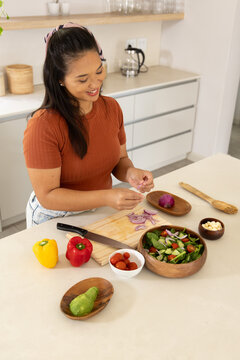 Mid-adult Asian woman preparing salad at kitchen counter in rust top and jeans using wooden board