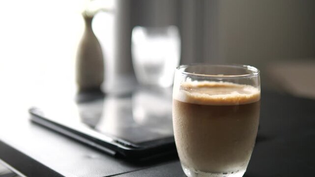 Close-up of iced latte coffee in a glass on a table with a blurred digital tablet and stylus, remote work and cafe lifestyle concept