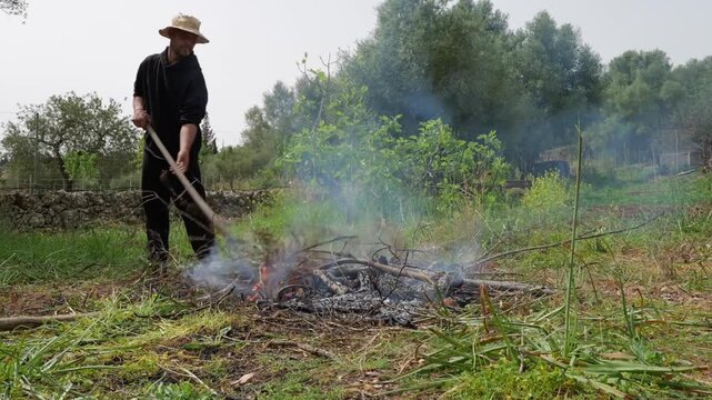 Man burning organic pruning waste in a rural field. Yellow wheelbarrow with logs in foreground. Small bonfire with smoke during seasonal garden maintenance and farm work.