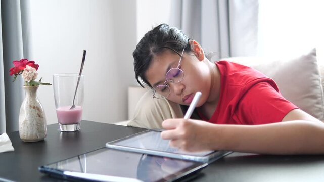 Close-up of a tired young student girl wearing glasses, leaning her head on her arm while using a digital tablet and stylus for online learning, exhausted and sleepy concept