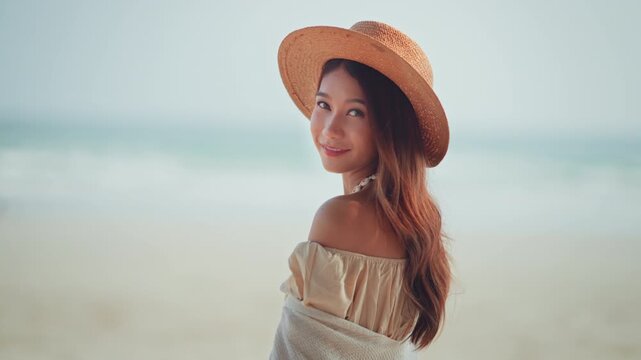 Beautiful Asian young woman wearing a straw hat smiling while looking back at the camera on a tropical beach during summer vacation. A happy female traveler enjoying a peaceful holiday and wellness