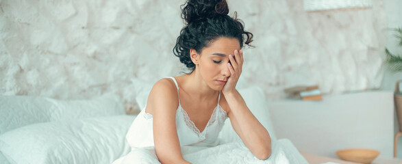 Naklejka premium A woman sits on her bed with her hand on her forehead. She appears to be in deep thought. The room is bright and has a minimalistic style. Natural light enters through the window.