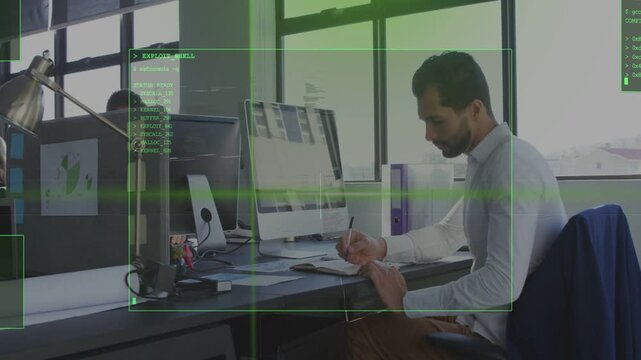 Man in tech office reviewing modules and writing notes while HUD overlay appearing around monitor